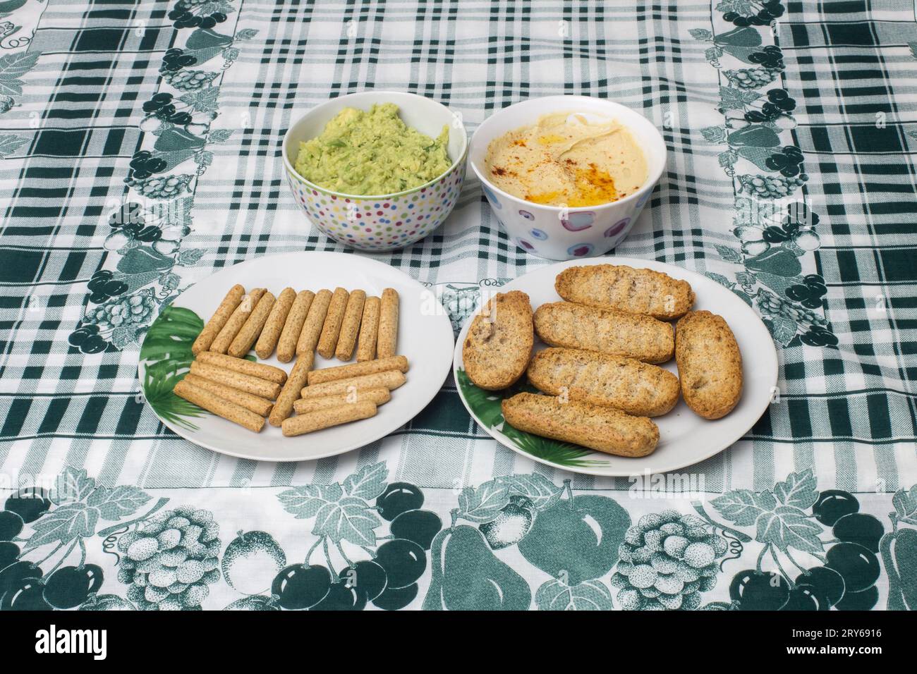 Zwei kleine Schüsseln mit hausgemachtem Hummus und Guacamole, begleitet von Platten mit Vollkorntoast und Toaststäbchen auf einer stilvollen Tischdecke Ador Stockfoto