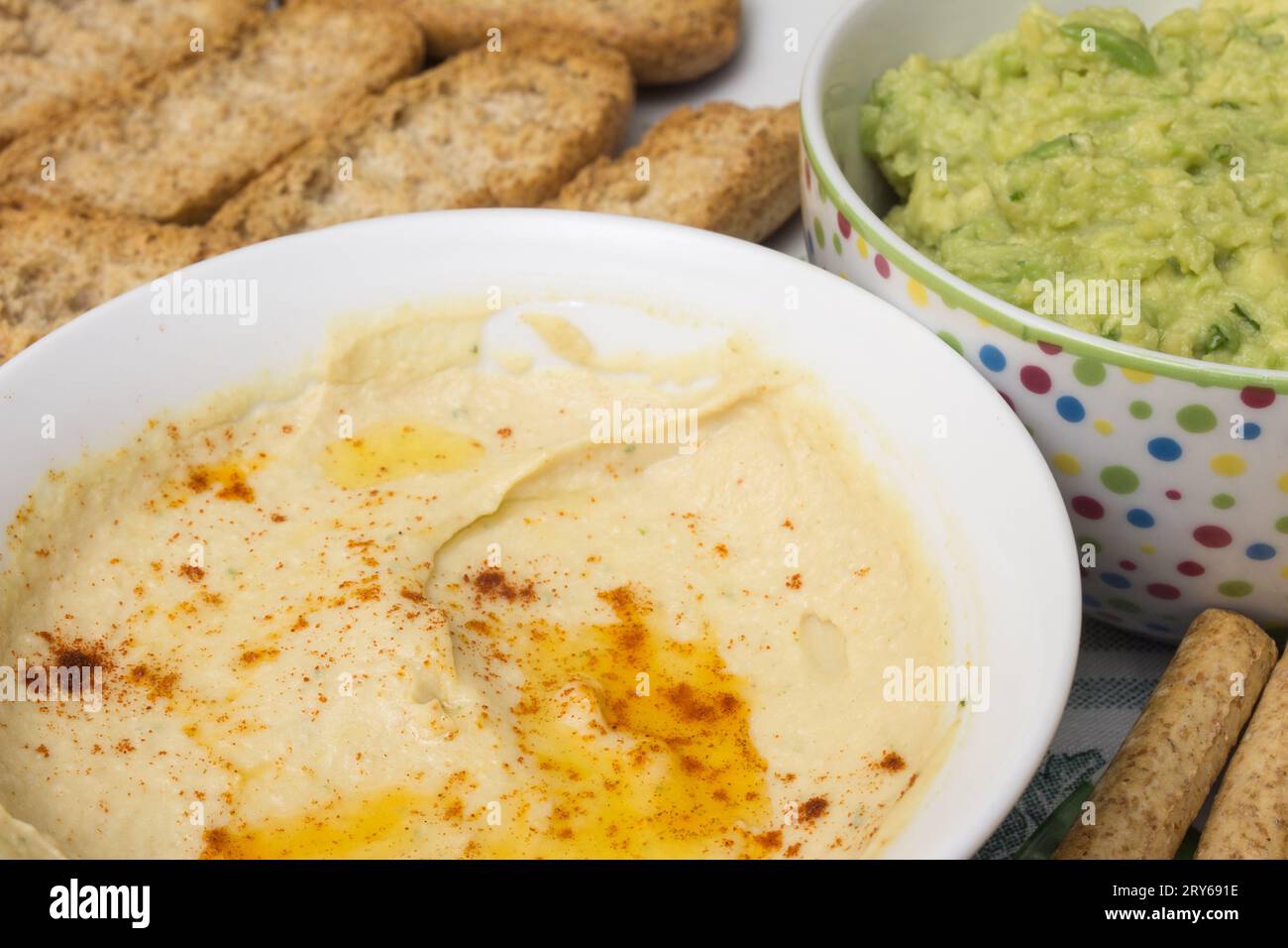 Eine Schüssel Hummus-Dip aus Kichererbsen, Tahini, Knoblauch und Zitrone. Der Hintergrund ist verschwommen, aber man sieht eine Schüssel Guacamole Stockfoto