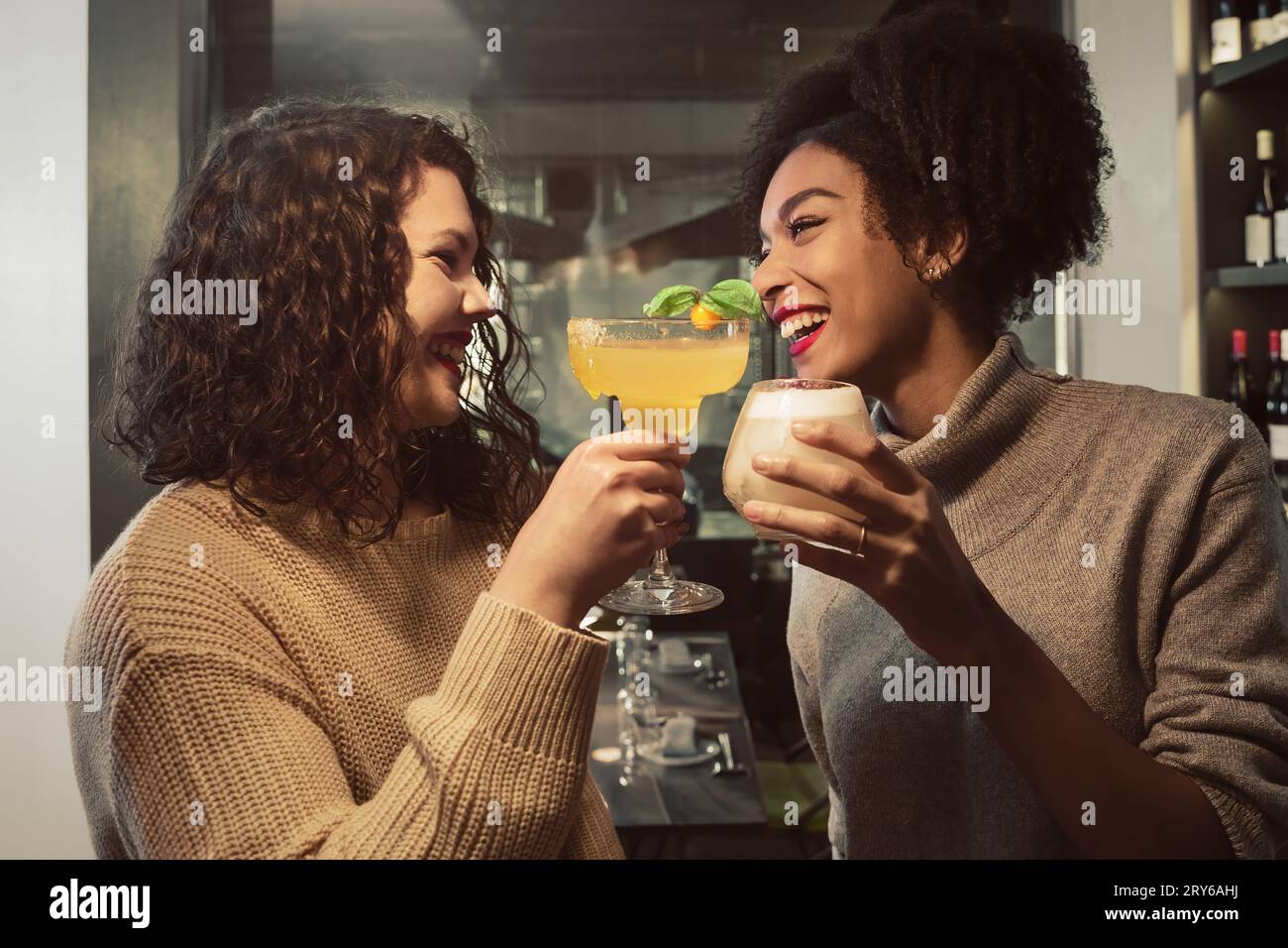 Zwei junge Frauen, eine Afroamerikanerin und eine Kaukasierin, schauen sich lachend in die Augen und toasten in einer Bar mit Cocktails. Beide tragen eine kuschelige Sweatjacke Stockfoto