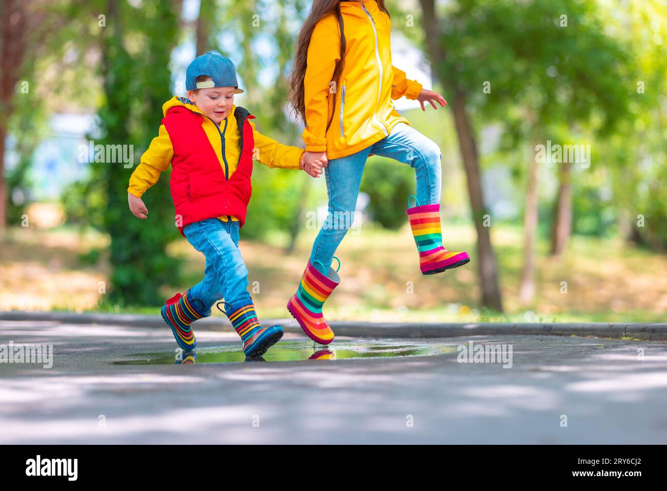 Glückliche Kinder, Mädchen und Junge mit Regenschirm und bunten Gummistiefeln, die draußen spielen und in regnerischer Pfütze springen Stockfoto