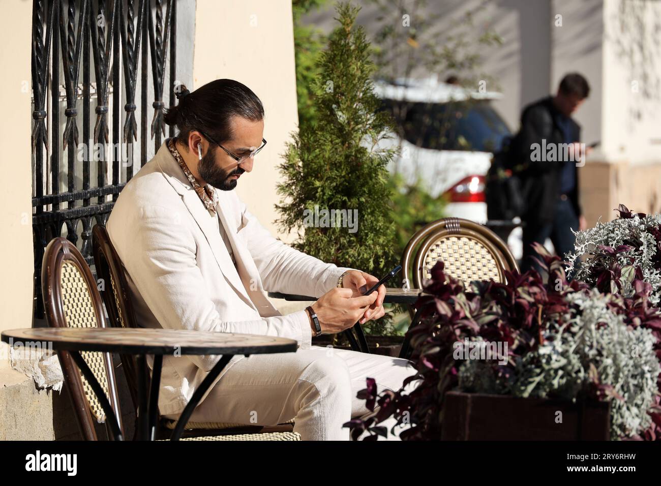 Gutaussehender bärtiger Mann in Brille und weißem Anzug, der mit Smartphone im Straßencafé sitzt Stockfoto