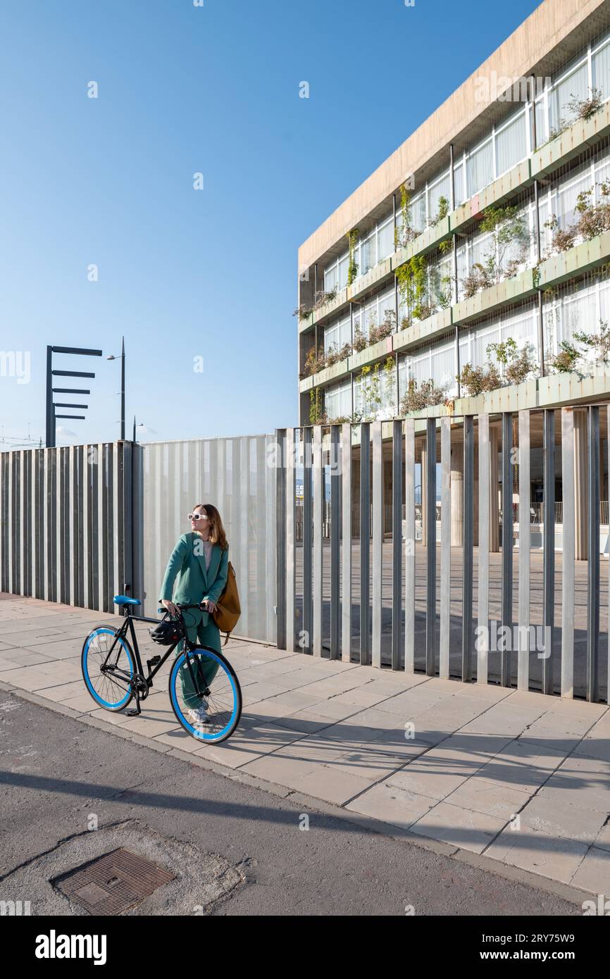 Stilvolle Geschäftsfrau im grünen Anzug, die mit dem Fahrrad im Finanzviertel mit modernem Gebäude im Hintergrund spazieren geht Stockfoto