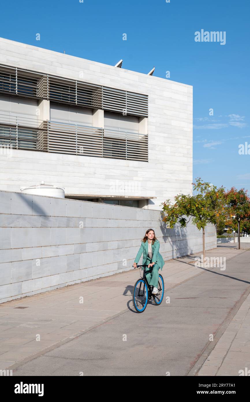 Stilvolle Geschäftsfrau im grünen Anzug, die im Finanzviertel mit modernem Gebäude im Hintergrund mit dem Fahrrad reitet Stockfoto