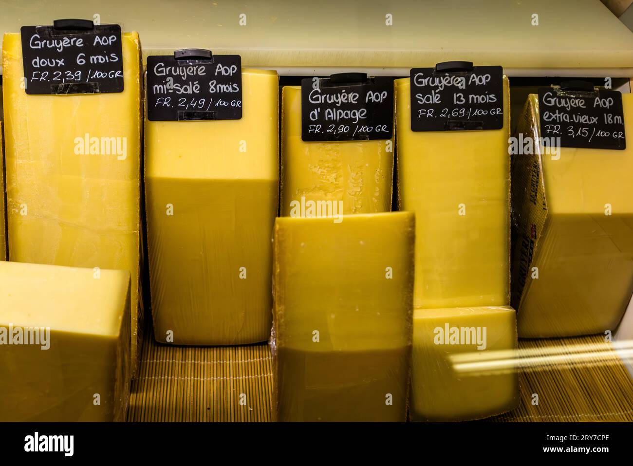 Mehrere Käsewürfel aus Gruyère AOP in verschiedenen Reifegraden liegen mit Preisschildern an einer Käsetheke in Freiburg, Schweiz. Zu den ausgestellten Sorten gehören „doux“ (6 Monate), „mi-salé“ (8 Monate), „d'alpage“, „salé“ (13 Monate) und „extra-vieux“ (18 Monate). Stockfoto