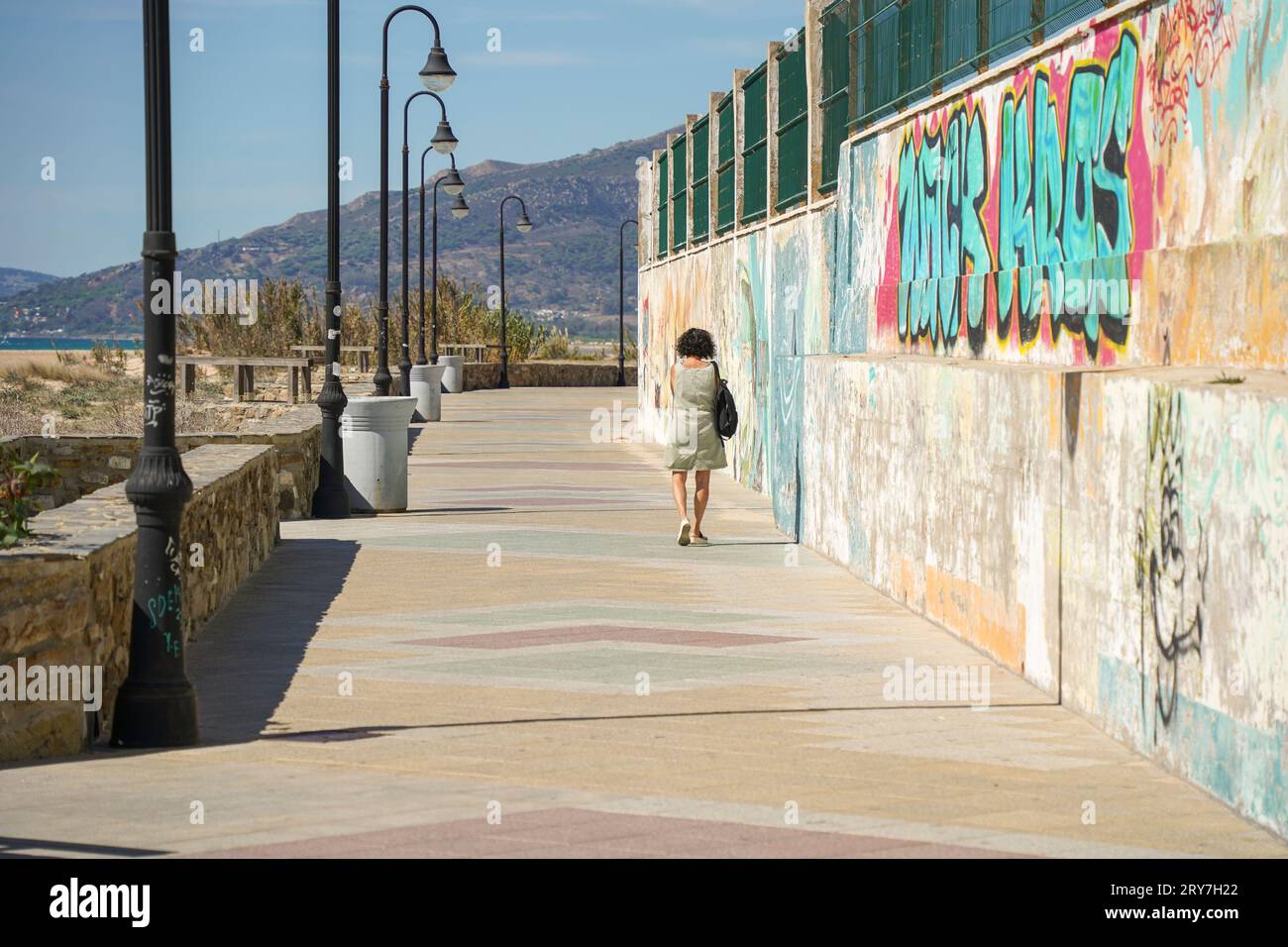 Frau geht allein, Fußgängerweg, Promenade mit Straßenlaternen und Graffiti an der Wand neben dem Strand, Tarifa, Spanien. Stockfoto