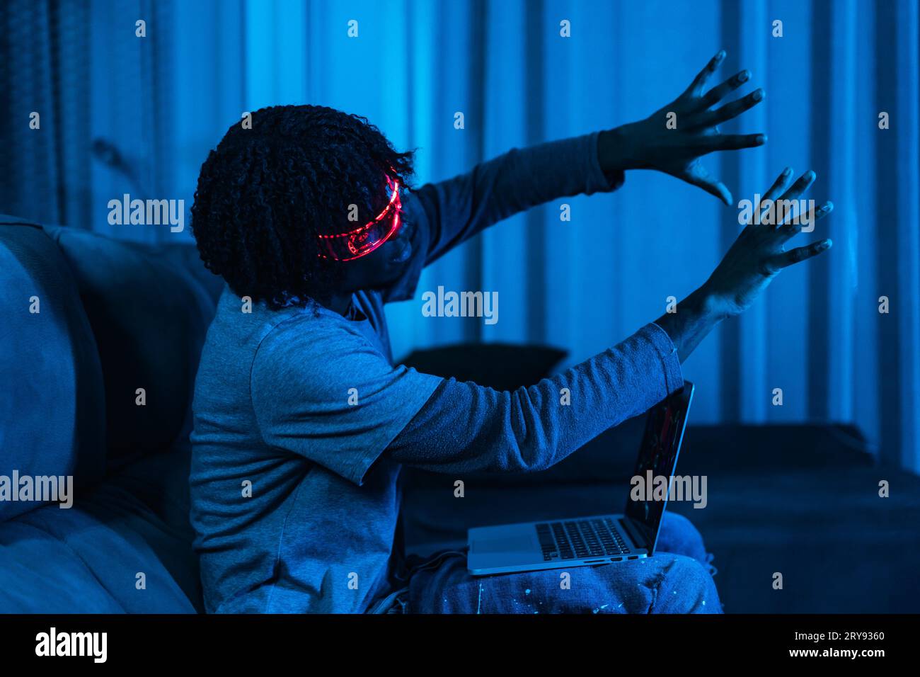 Afro man spielt mit Laptop und Augmented Reality Brille in einem Wohnzimmer mit Neonblau Licht Stockfoto