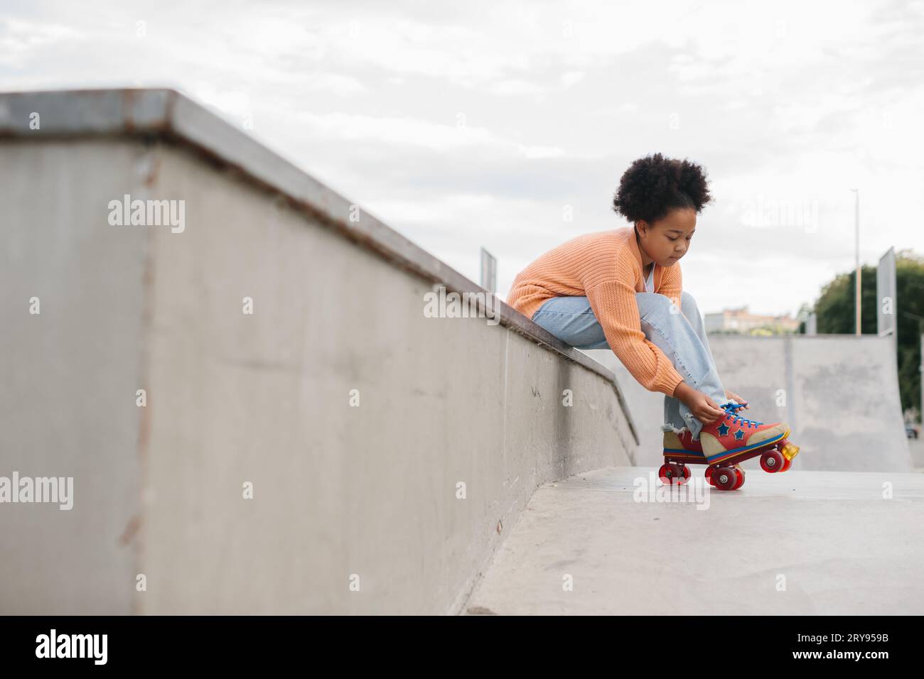 Süßes afroamerikanisches Teenie-Mädchen, das Rollschuhe anzieht, während es auf einer Rampe in einem Skatepark am Strand sitzt. Stockfoto