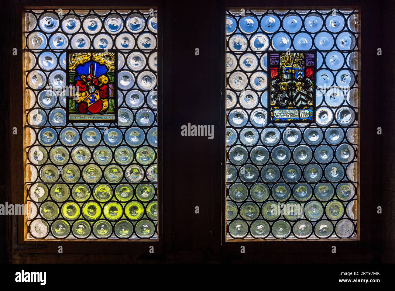 Zwei runde Bullauge-Fenster mit Wappen in Glasmalerei erhellen das, was wahrscheinlich der Scharfrichter-Saal (Salle des baillis) im Schloss Gruyères in Épagny bei Gruyères, Kanton Freiburg, Schweiz, ist. Recherchen deuten darauf hin, dass die farbigen Wappenfenster aus dem Jahr 1685 stammen, wie auch der Rest der Einrichtung des Saals. Das im 13. Jahrhundert erbaute Schloss war bis 1554 der Sitz der Grafen von Gruyère. Seit 1938 gehört es dem Kanton Freiburg und dient als Museum. Stockfoto