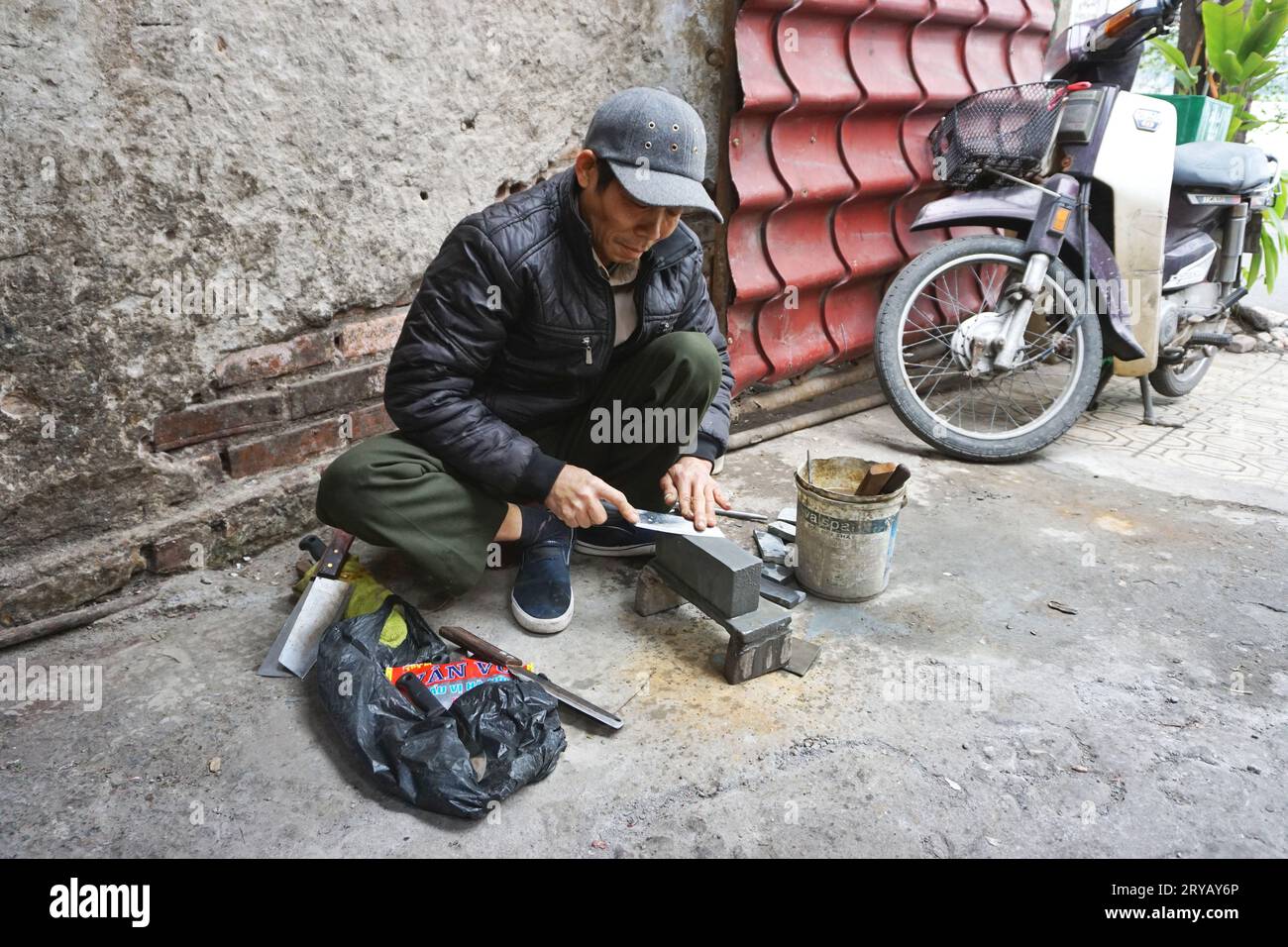 Ein reisender Messerschärfer macht seinen Beruf außerhalb eines traditionellen Feuchtmarktes in Hanoi, Vietnam. Die Fleischverkäufer dort sind seine Stammkunden. Stockfoto