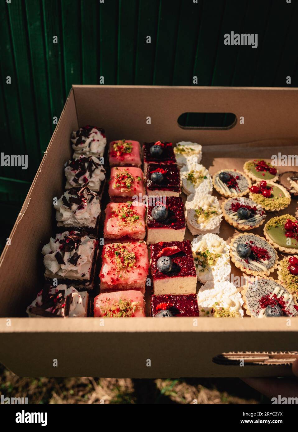 Nahaufnahme eines Papierkartons voller Kuchen und Torten (Punschkuchen, Mini pavlova usw.) auf grünem Hintergrund. Baker (Frau) hält ihre Schachtel mit leckerem und bea Stockfoto