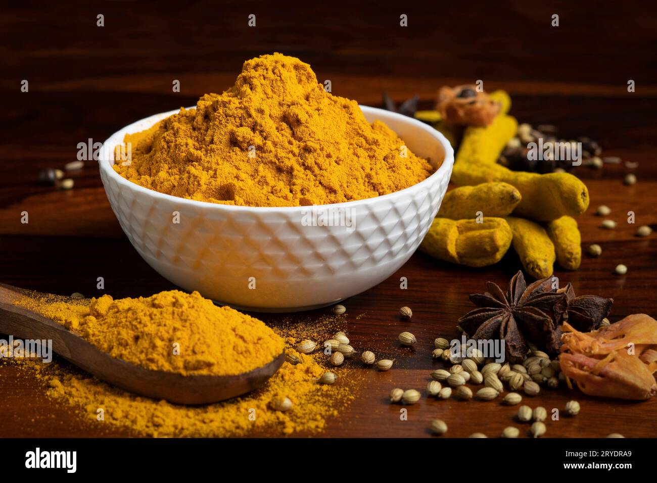 Eine Schüssel mit gelbem Kurkuma-Pulver, umgeben von einem Holzlöffel mit Kurkuma-Pulver, trockenen Gewürzen wie Sternanis, Muskatnuss, Kümmel, Koriandersamen, trocken Stockfoto
