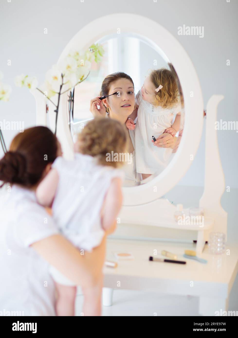 Mutter und Tochter blicken in den Spiegel. Junge Mutter und kleines Mädchen tragen Make-up auf und bürsten Haare. Schöne Frau mit entzückendem Baby im weißen Schlafzimmer Stockfoto