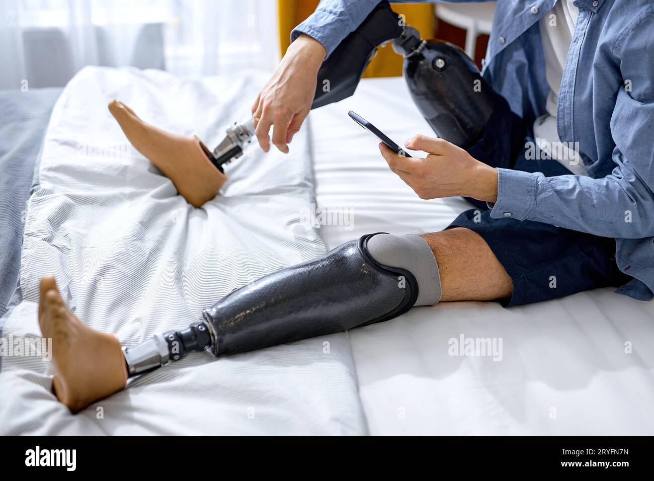 Nicht erkennbarer behinderter Mann mit künstlichem Bein bleibt zu Hause. Erwachsener Mann mit Beinprothesen im Ruhezustand. Mann in lässiger Haushaltskleidung sitzt auf dem Bett h Stockfoto