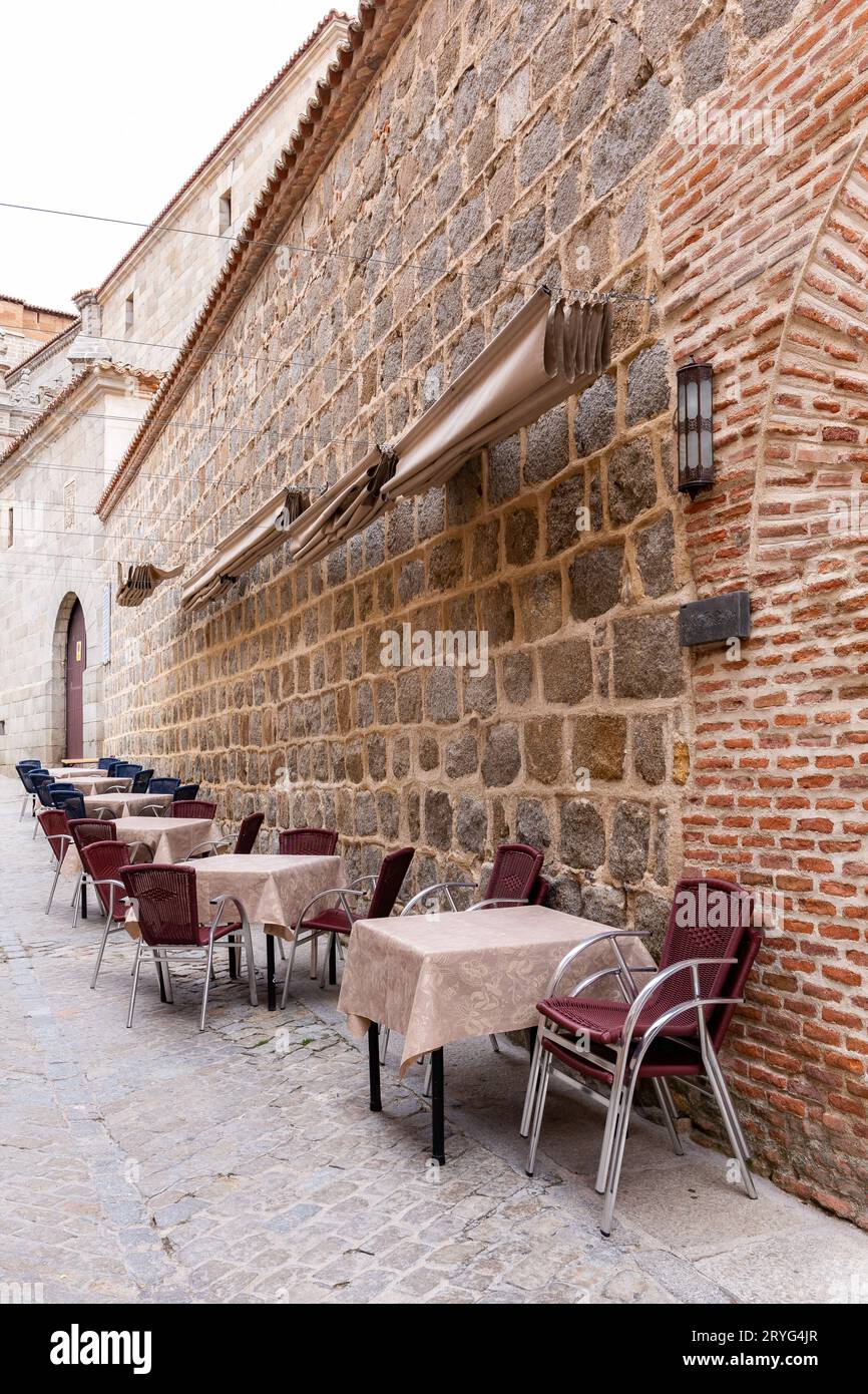 Mittelalterliche, schmale Kopfsteinpflasterstraße in Avila, Spanien, mit Restauranttischen und Essbereich, keine Leute. Stockfoto