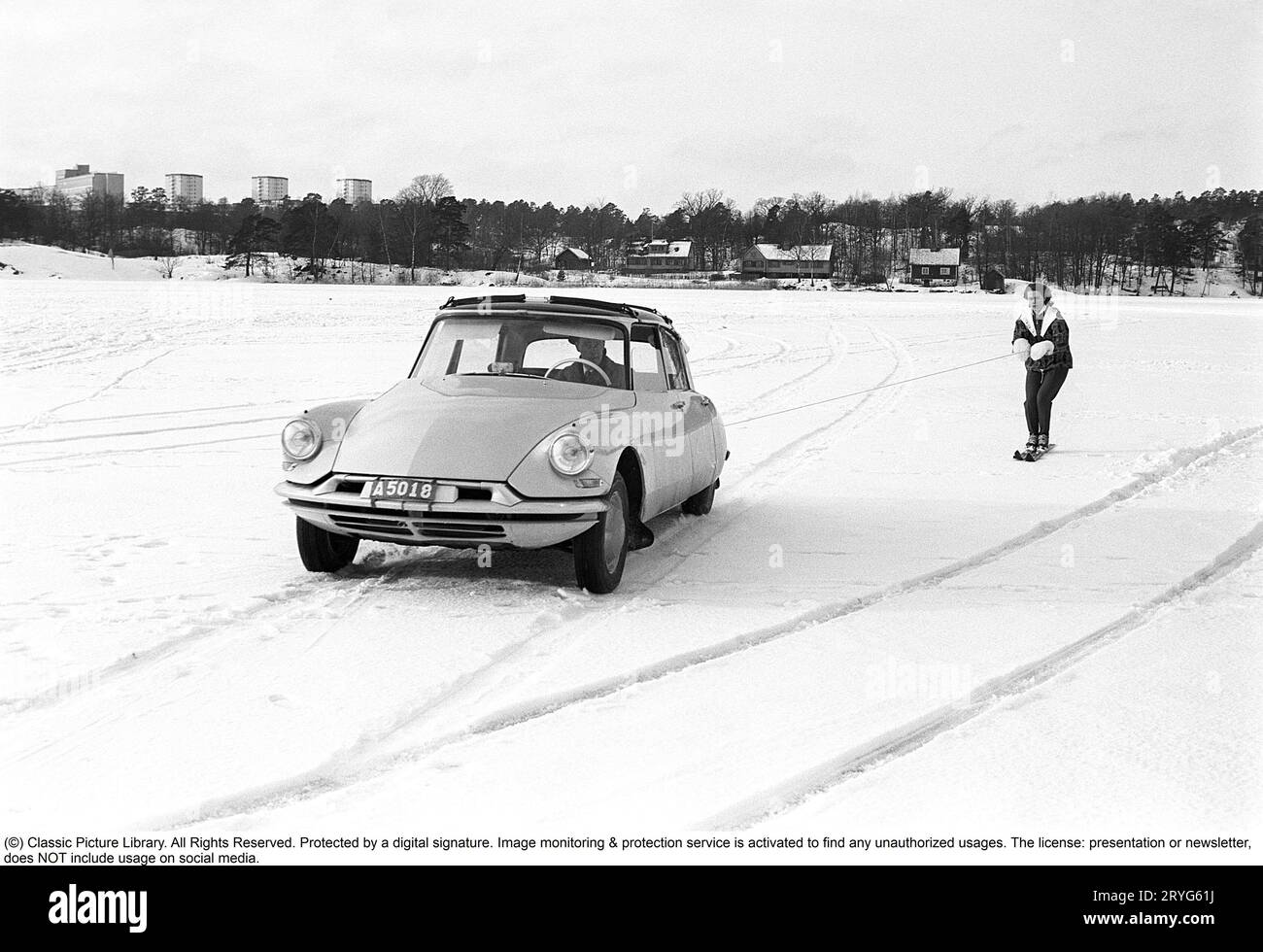 Ein Wintertag in den 1960er Jahren Eine Frau fährt hinter einem Auto. A Citroën. Es muss ziemlich dickes Eis auf dem See gewesen sein, weil der Fahrer es wagte, auf dem Eis zu fahren, aber selbst die Frau muss mutig gewesen sein, als sie sicher Geschwindigkeit auf ihren Skiern nahm, als sie an einem Seil mit einem Griff hinter dem Auto an hielt volle Geschwindigkeit. Schweden 1962. Conard Ref. 4561 Stockfoto
