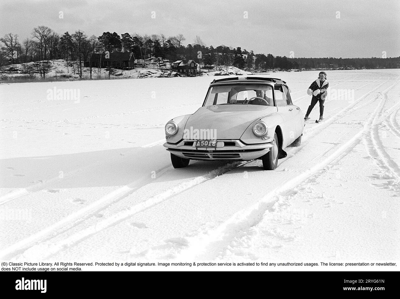 Ein Wintertag in den 1960er Jahren Eine Frau fährt hinter einem Auto. A Citroën. Es muss ziemlich dickes Eis auf dem See gewesen sein, weil der Fahrer es wagte, auf dem Eis zu fahren, aber selbst die Frau muss mutig gewesen sein, als sie sicher Geschwindigkeit auf ihren Skiern nahm, als sie an einem Seil mit einem Griff hinter dem Auto an hielt volle Geschwindigkeit. Schweden 1962. Conard Ref. 4561 Stockfoto