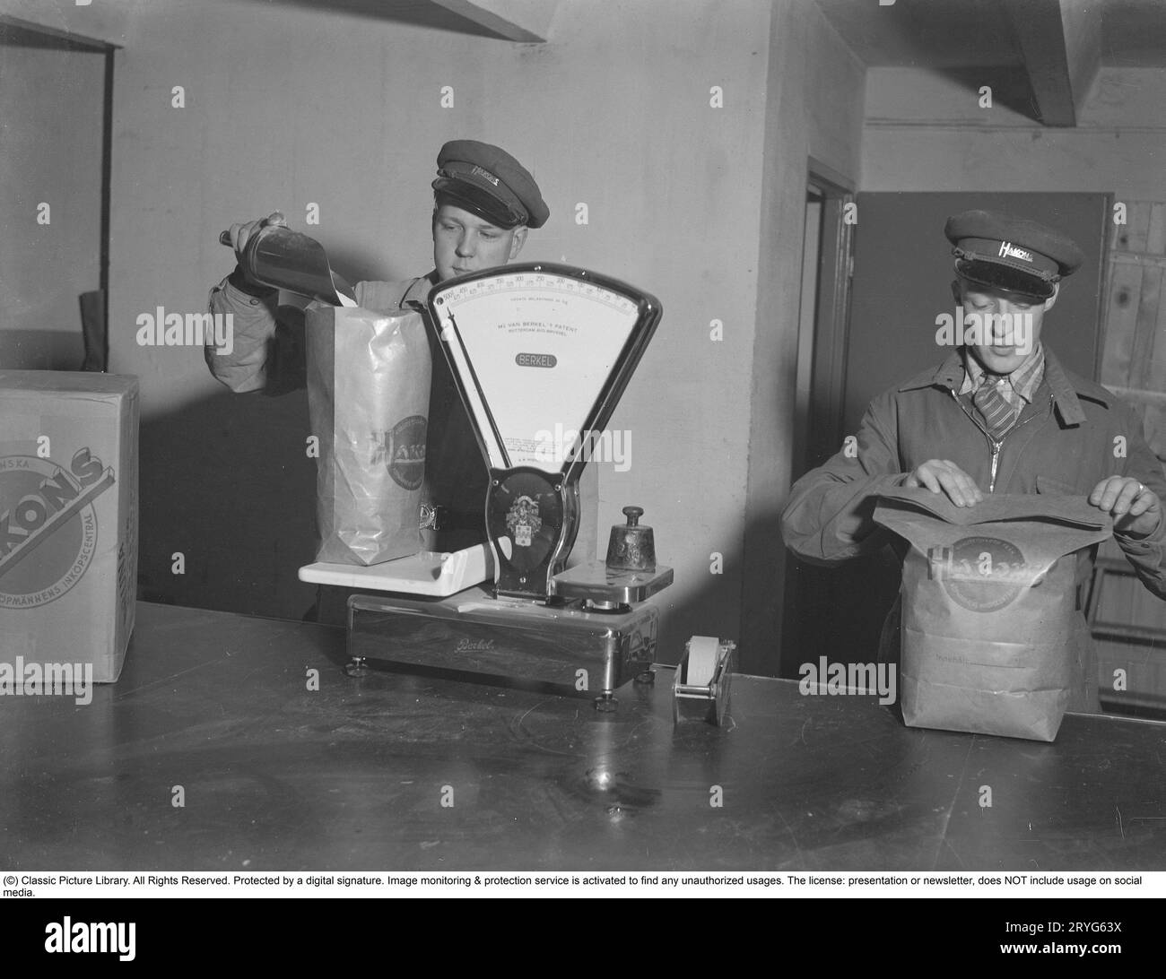 In den 1940er Jahren Lars Nilsson wiegt Mehl in einem Beutel auf einer Waage. Auf den Taschen steht das Einkaufszentrum von Hakon Köpmännen. Die Firma Hakon heißt jetzt ICA Hakon. Eine Unternehmensgruppe, die heute eine der größten Lebensmittelketten Schwedens ist. Lars Nilsson war ein schwedischer Leichtathlet und gewann 1940 die erste kleine Goldmedaille im Langlauf. Kristoffersson, Ref. 123-7 Stockfoto