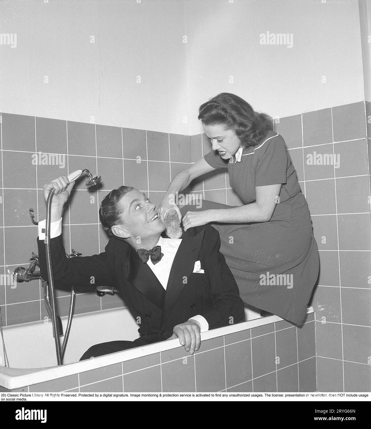 Seltsam in den 1940er Jahren Sture Lagerwall, 1908-1964. Schwedische Schauspielerin hier mit Schauspielerin Sonja Wigert, die beide in einer Badewanne bekleidet gesehen werden. Er hält die Dusche über sich, ohne Wasser auf sich zu spritzen, und die Frau wäscht ihn verspielt mit einer Bürste. Schweden 1945. Kristoffersson R47-6 Stockfoto