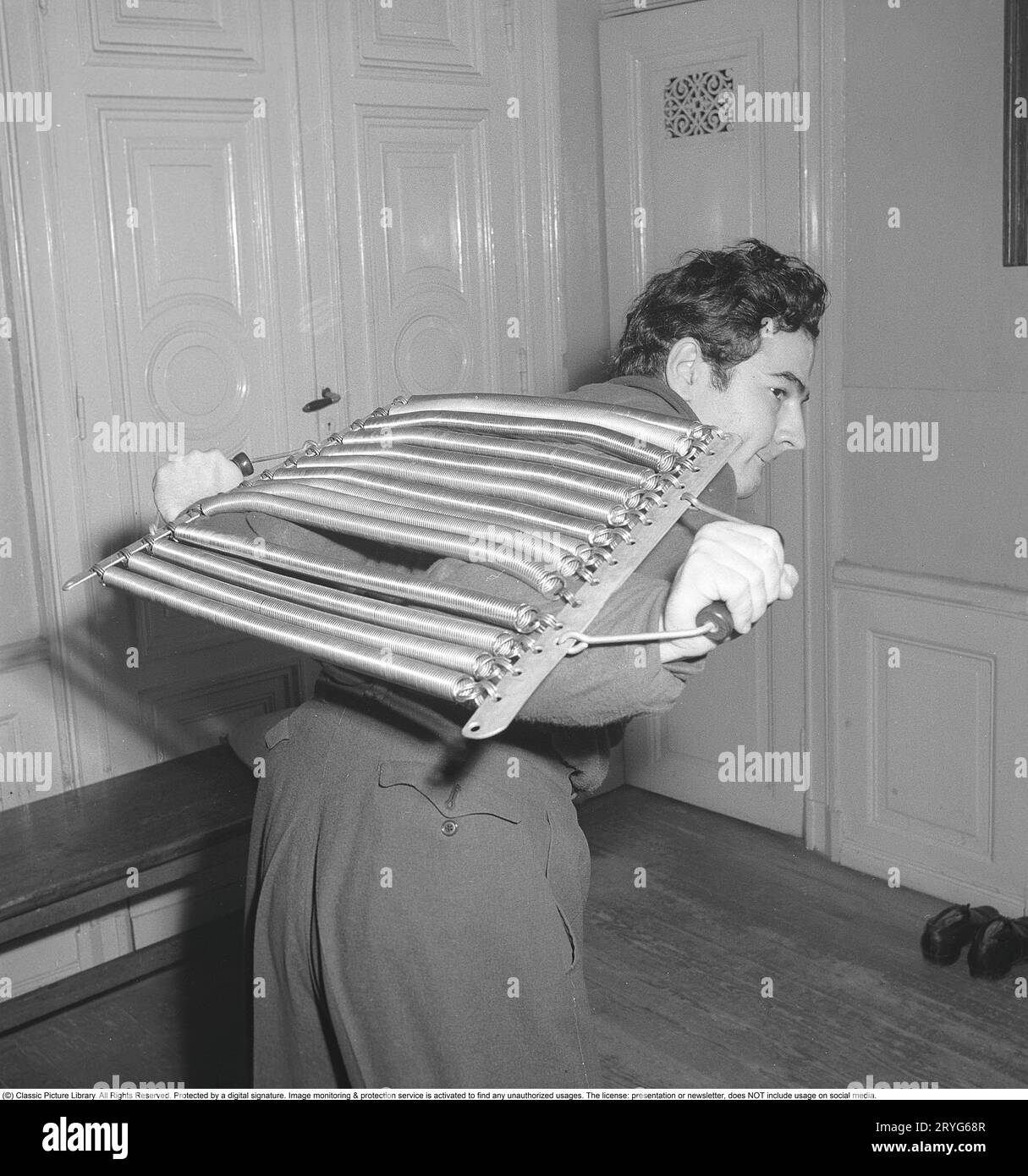 Krafttraining in den 1940er Jahren Ein junger Mann trainiert seine Muskeln mit einem großartigen Trainingsgerät. Er zieht die vielen Federn in den Trainingsgeräten auseinander, die sicher für viel Widerstand sorgen und auf diese Weise seine Muskeln stärken und trainieren. Schweden 1946. Kristoffersson ref X110-5 Stockfoto