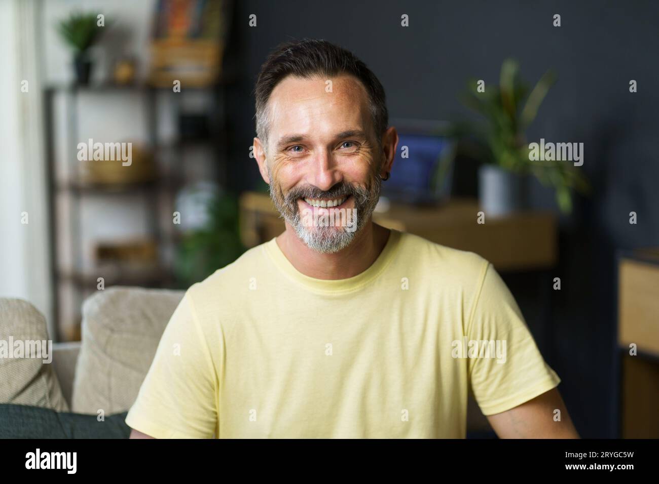 Porträt eines gutaussehenden, fröhlichen grauhaarigen freiberuflichen Mannes mittleren Alters, der auf dem Sofa sitzt und die Kamera in lässiger Kleidung ansieht. Bedacht Stockfoto