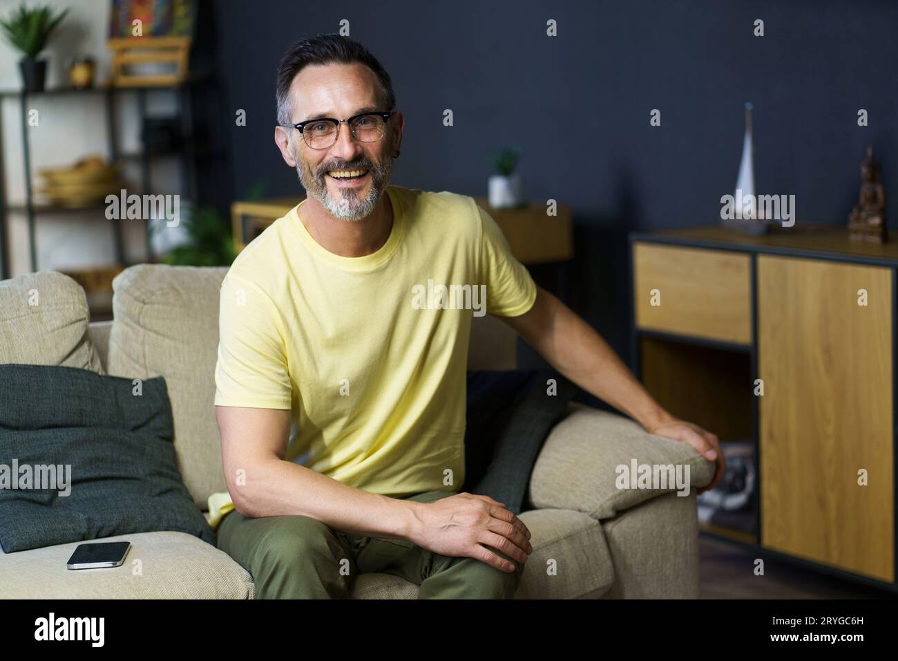 Fröhlicher grauhaariger Mann mittleren Alters, der auf dem Sofa sitzt und eine Brille trägt und die Kamera in lässiger Form betrachtet. Rücksichtsvoller Freiberufler Stockfoto