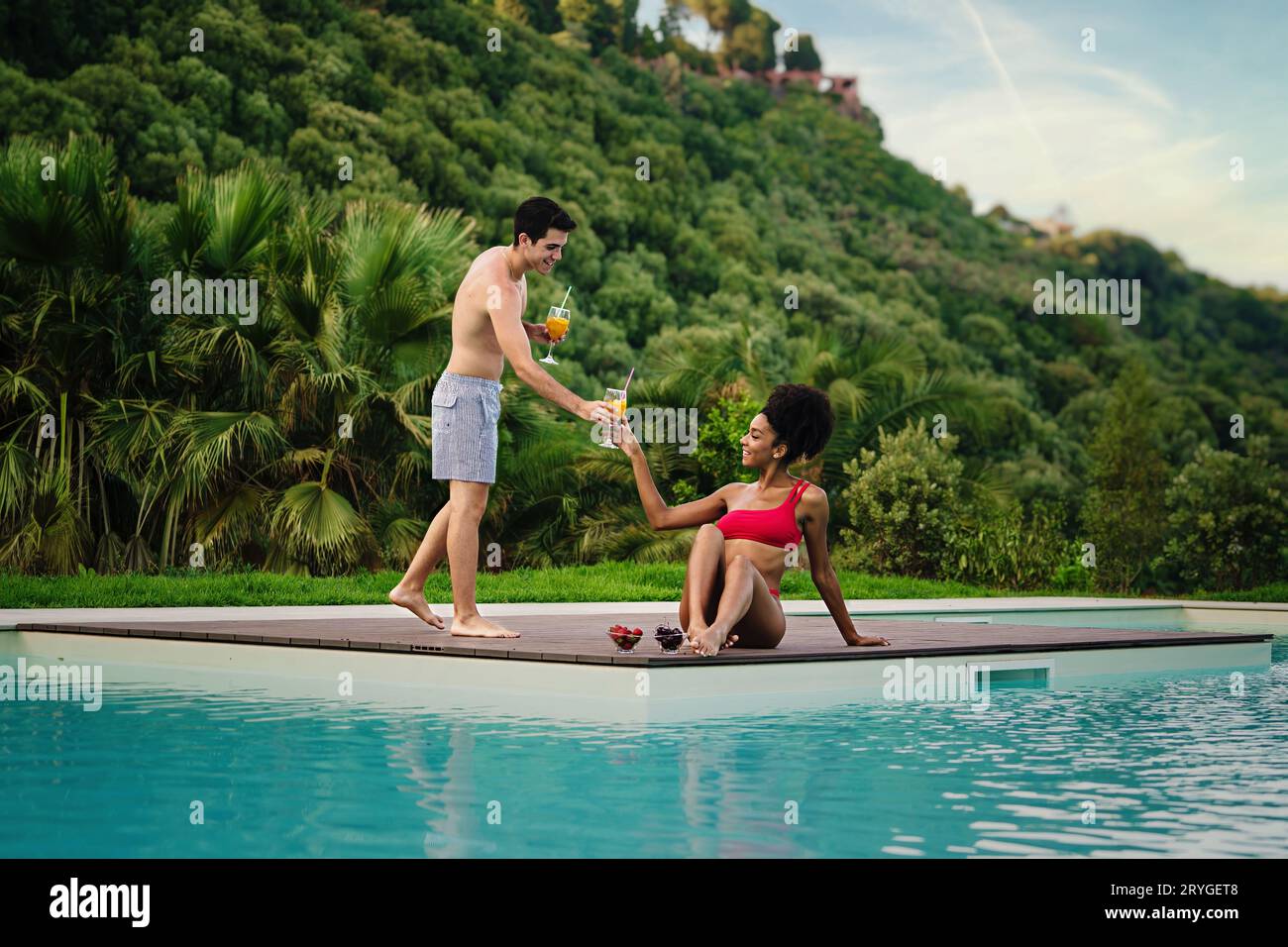 Ein Paar, das den Sommernachmittag am Pool genießt: Kaukasischer Mann in bermuda-Shorts, der seiner afroamerikanischen Freundin Orangensaft in einer Schulter anbietet Stockfoto