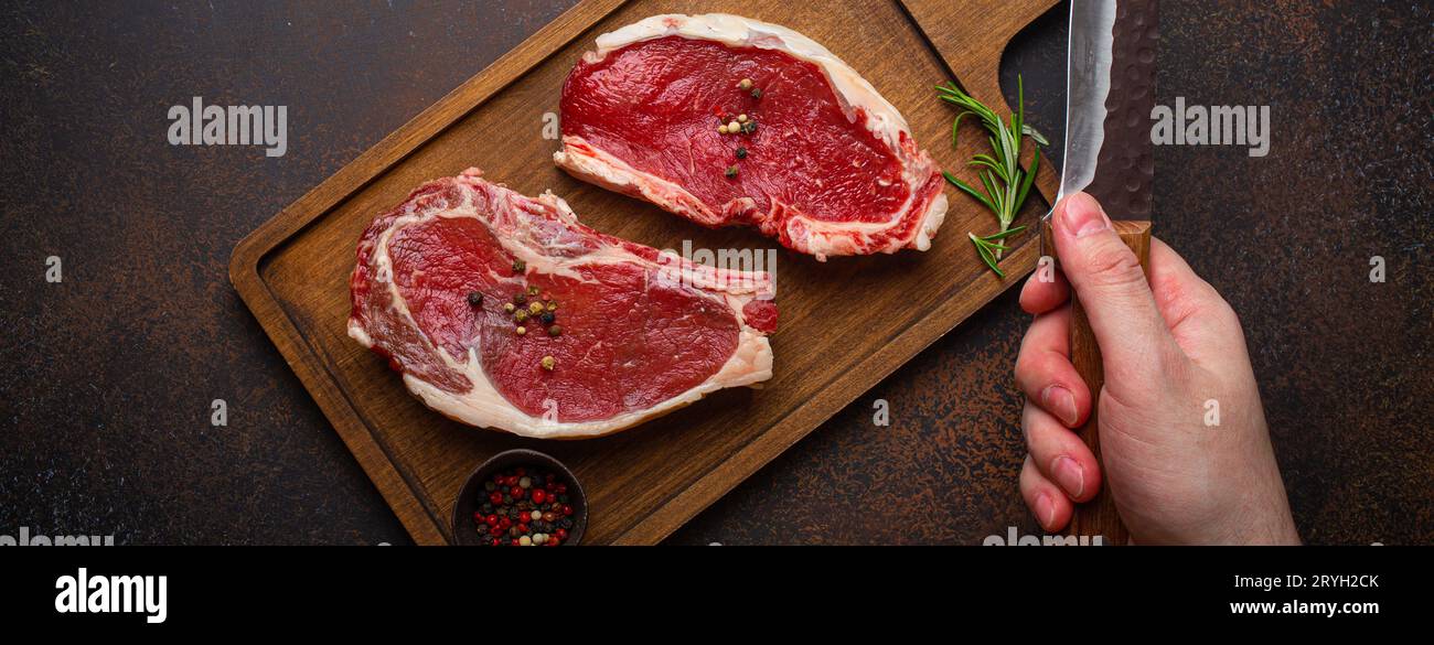 Männliche Hand hält Messer über zwei rohen Fleisch Rindfleisch Steaks Draufsicht Stockfoto