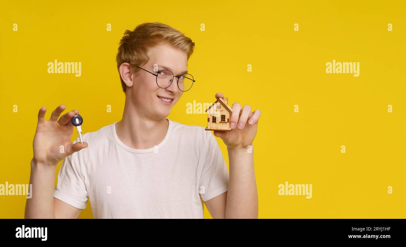 Junger Mann, der ein Architekturmodell, eine Maquette und einen Schlüssel vor gelbem Hintergrund hält, zeigt die Leistung des eigenen Hauses Stockfoto