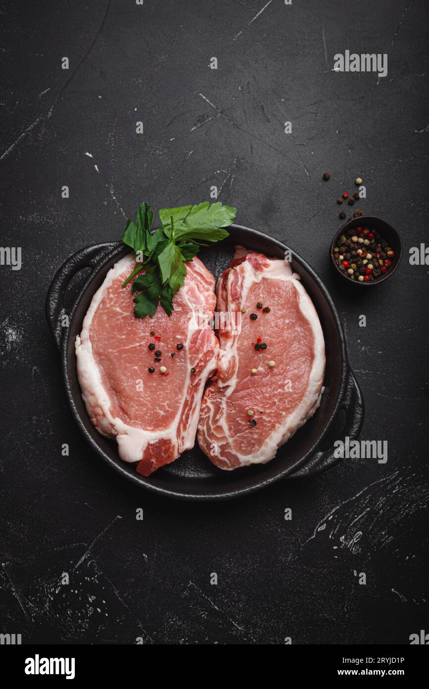 Schneiden Sie rohe Schweinefleisch-Steaks mit Gewürzen in einer schwarzen gusseisernen Pfanne auf dunklem rustikalem Steinhintergrund von oben, bereit zum Rösten. Po Stockfoto