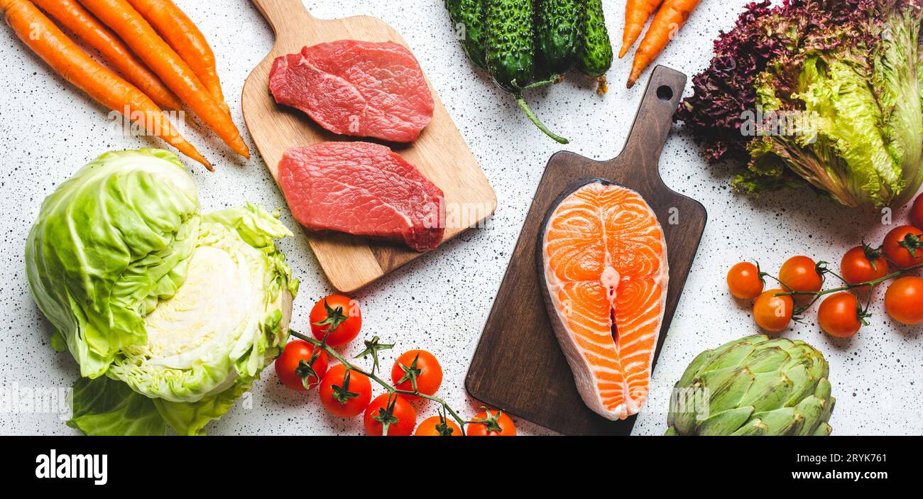Verschiedene rohe Lebensmittel: Gemüse, Rindfleischsteak, Fisch Lachsfilet auf hölzernen Schneidebrettern, weiße rustikale Tischsicht Stockfoto
