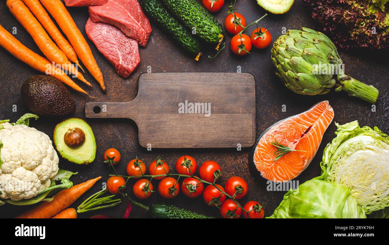 Verschiedene Lebensmittel-Rohprodukte: Gemüse, Rindfleisch, Fischlachs und leeres Holzschneidebrett in der Mitte, dunkler rustikaler brauner Stein Stockfoto