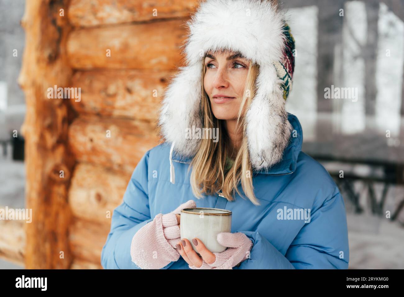Porträt einer kaukasischen Frau in einem Winterhut mit Ohrenklappen mit einem Becher heißen Getränks. Stockfoto