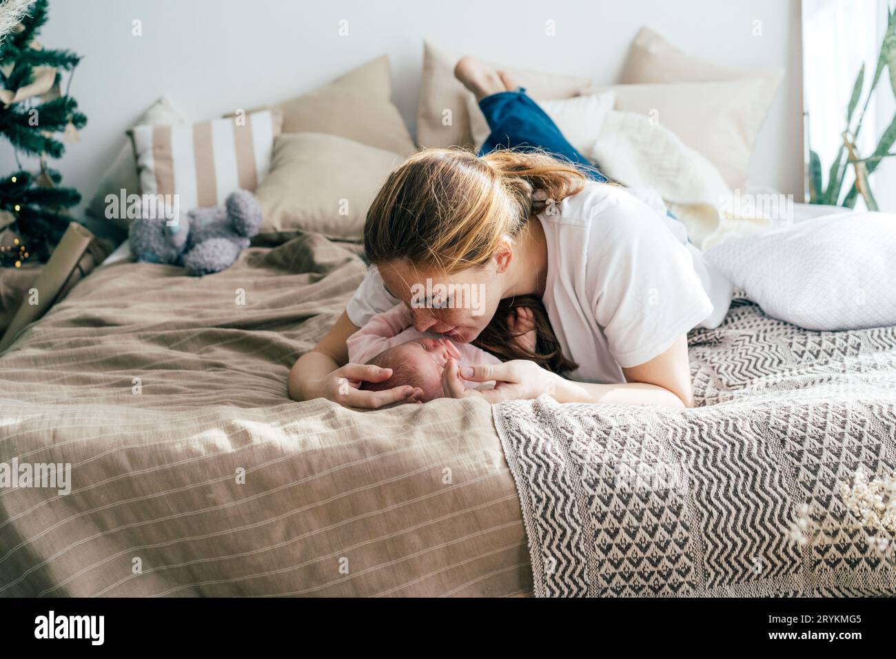 Eine zarte Mutter, die auf dem Bett liegt, umarmt und küsst das Baby. Authentisches Familienportrait. Mutterschaft. Stockfoto