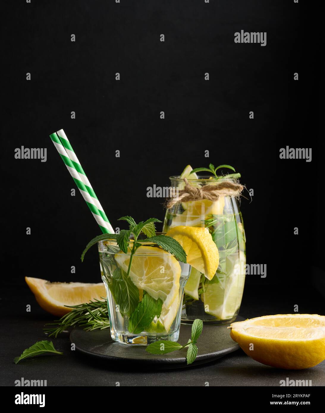 Limonade in einem transparenten Glas mit Zitronen-, Limonen-, Rosmarin-Zweigen und Minzblättern auf schwarzem Hintergrund Stockfoto