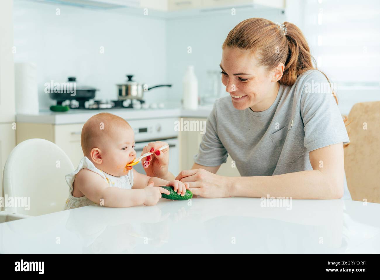 Glückliche, lächelnde rothaarige Mutter füttert ihr Baby mit Gemüsepüree in der Küche zu Hause. Tagesablauf der Kinderbetreuung und Erziehung Stockfoto