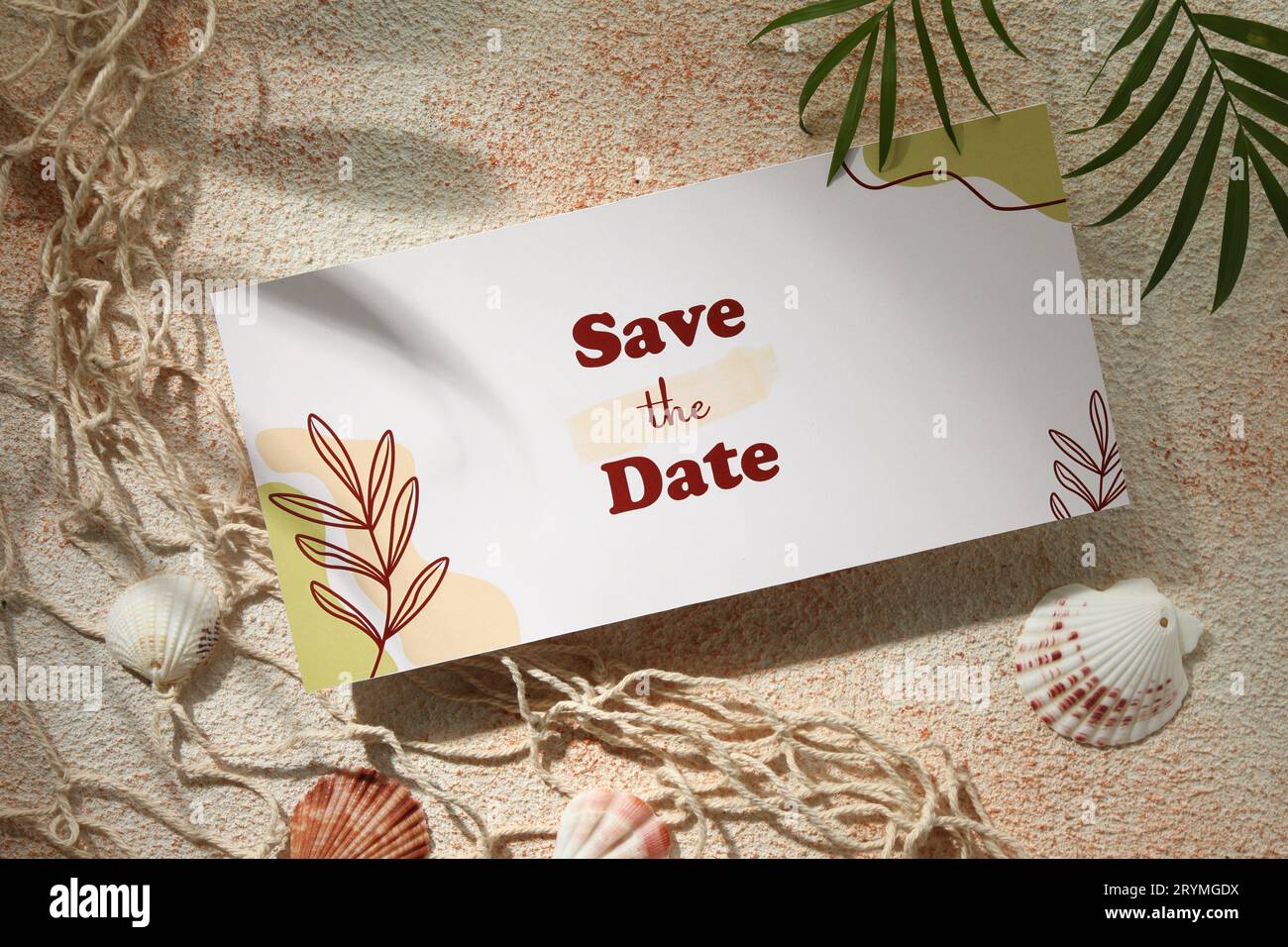 Schöne Hochzeitseinladungskarte mit „Save the Date“-Phrase, Muscheln, Netz und grünen Blättern auf strukturiertem beigem Hintergrund, flach liegend Stockfoto