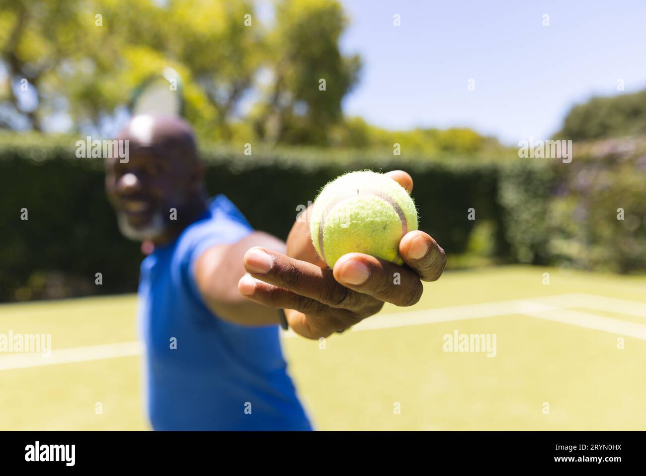 Glücklicher afroamerikanischer Senior-Mann, der Tennis spielt, Ball hält, bevor er auf dem sonnigen Rasenplatz serviert. Seniorenleben, Ruhestand, Sport, Sommer, Fitness Stockfoto