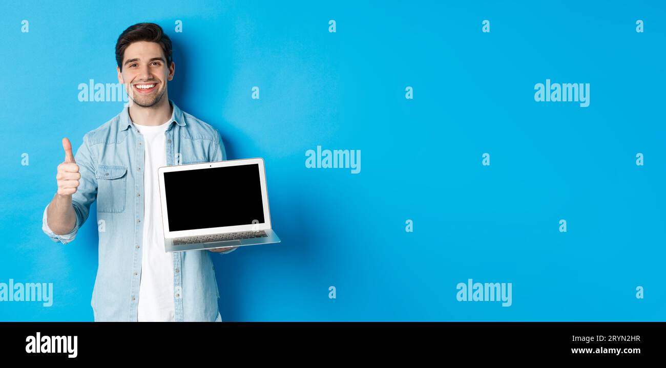 Ein lächelnder, gutaussehender Mann zeigt den Laptop-Bildschirm und den Daumen nach oben, wie ein Werbeangebot, empfahl die Website, steht vor blauem Hintergrund Stockfoto