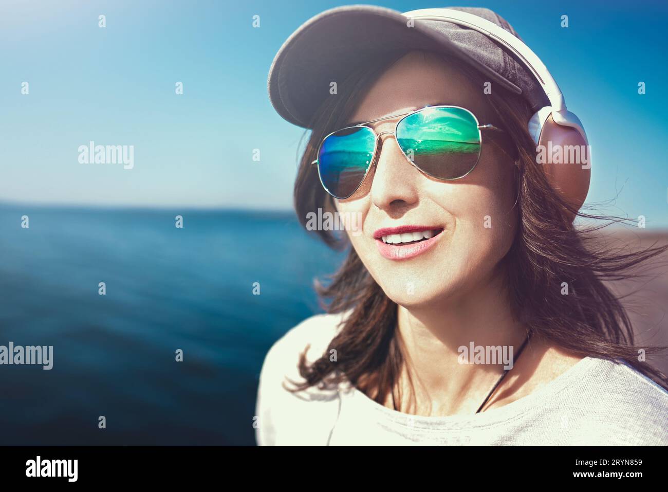 Nahporträt einer Frau in Sonnenbrille mit Mütze, die Musik im Hintergrund des Meeres genießt.Musikkonzept. Stockfoto