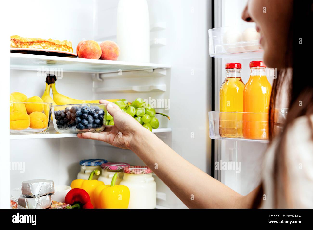 Frau, die eine Schachtel Blaubeere aus dem offenen Kühlschrankregal oder der Kühlschublade mit Obst, Gemüse, Bananen, Pfirsichen nimmt, schnappt oder nimmt, Stockfoto