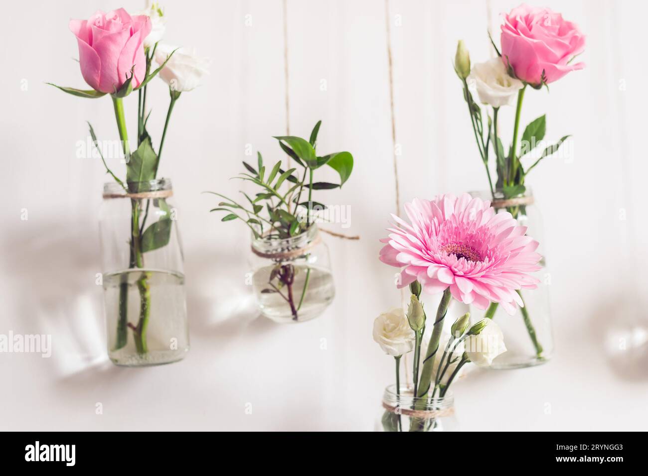 Schöne, stilvolle Urlaubsdekoration. Rosen und Gerbera in Gläsern hängen an der weißen Wand, Vorderansicht Stockfoto