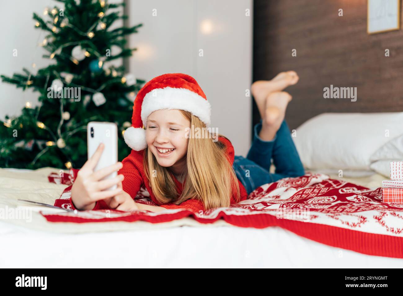 Ein wunderschönes, rothaariges Teenager-Mädchen in einem Weihnachtsmann-Hut, der auf dem Bett liegt, spricht bei einem Videochat am Telefon. Stockfoto