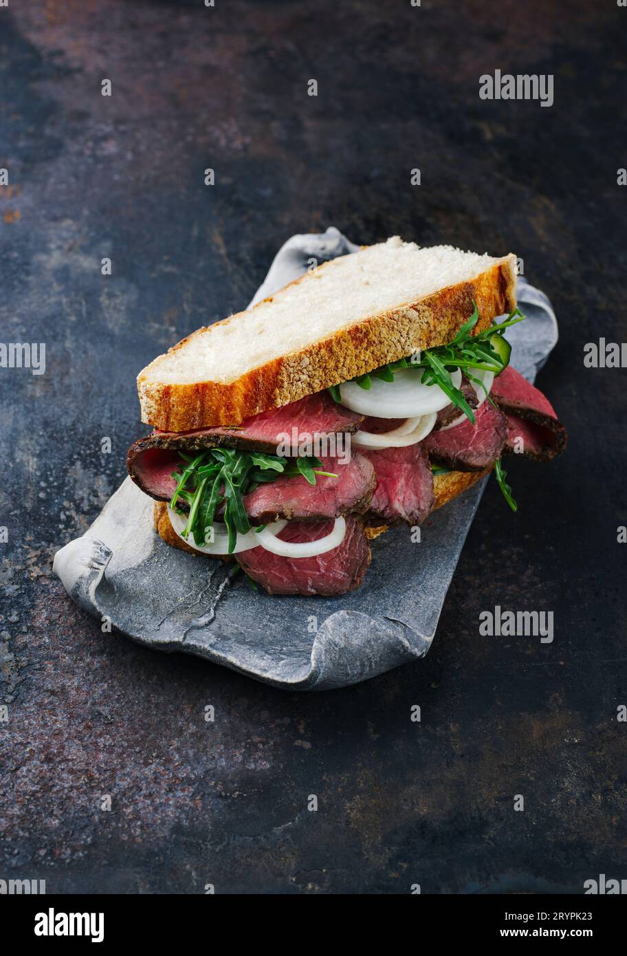 Traditionelles Roastbeef-Sandwich mit Raketensalat und Zwiebelringen, serviert mit italienischem Ciabatta-Brot als Nahaufnahme auf einem Design t Stockfoto