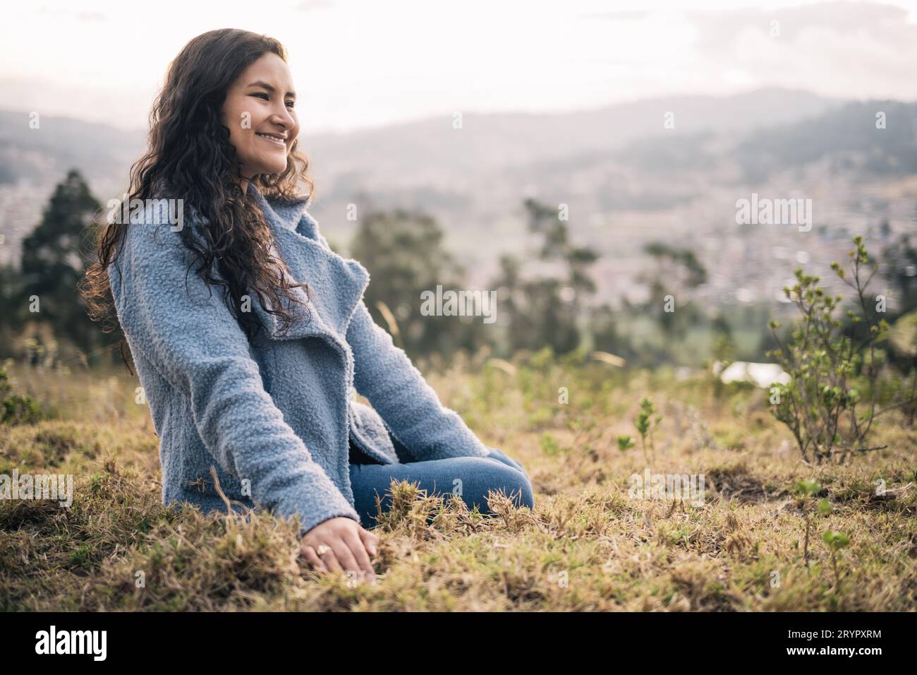 Eine schöne Lateinfrau, die auf dem Gras sitzt, lächelt draußen bei Sonnenuntergang Stockfoto