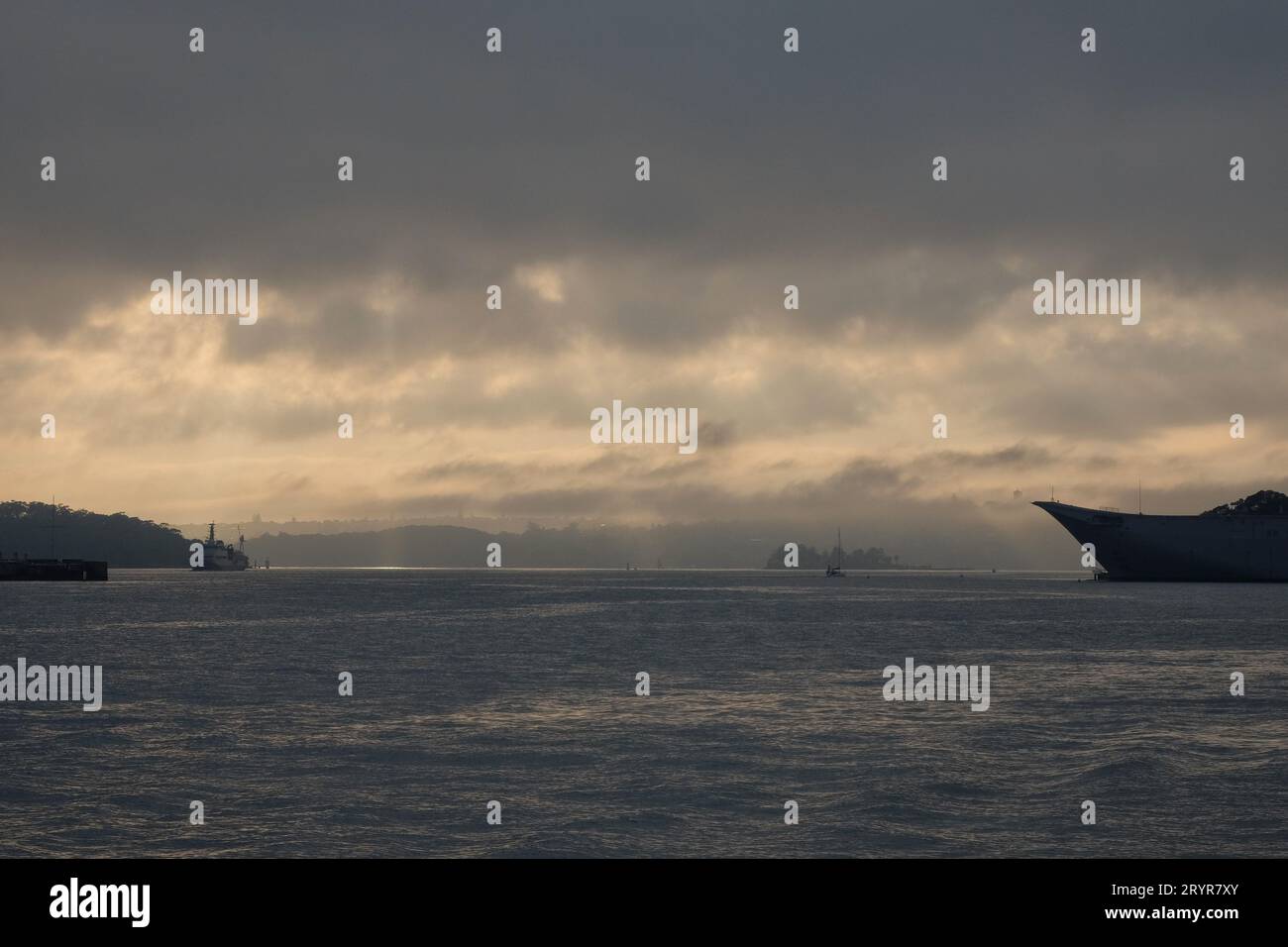 Die beeindruckende Schönheit des Hafens von Sydney, wenn Sonnenlicht durch Ufer niedriger Wolken bricht, Wasser kräuselt, winzige Boote, ein Schiffsbogen, Landstreifen Stockfoto