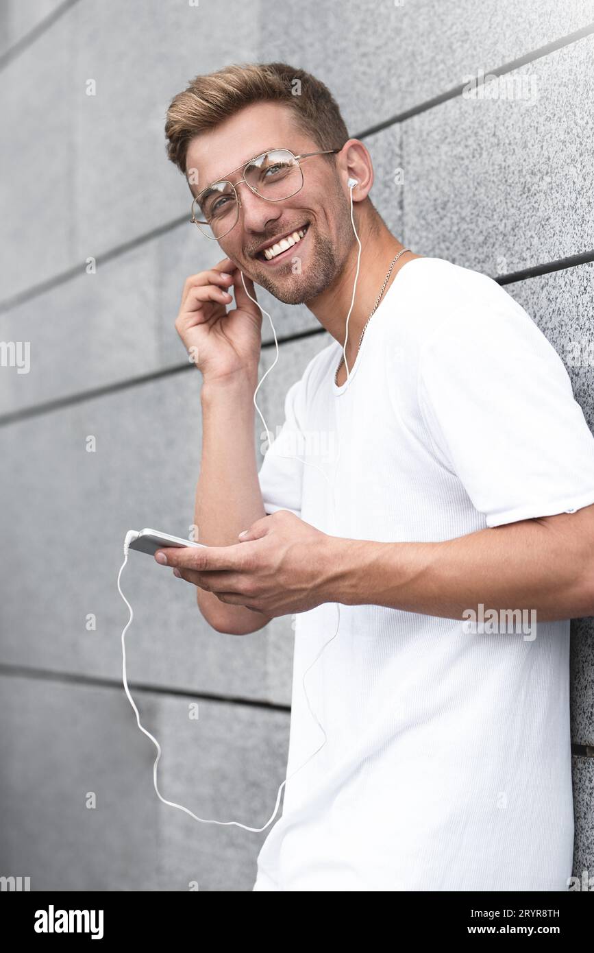 Fröhlicher Kerl in weißem T-Shirt auf der Straße, Musik mit Ohrhörern hören, Handy halten. Stockfoto