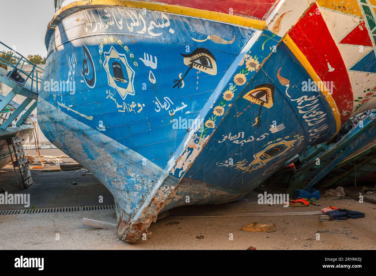 Alte Fischerboote, die von Flüchtlingen und Migranten genutzt wurden, um das Mittelmeer von Nordafrika nach Europa zu überqueren, von italienischen Grenzbeamten beschlagnahmt und im Hafen von Pozzallo, Sizilien, Italien, gelagert. Die gemalten Symbole (insbesondere das Horusauge) sollen Glück bringen und das Böse abwehren. Dieses Boot hat auch ein Bild der Ka'aba (in Mekka), die heiligste Stätte im Islam, sowie eine „Hamsa“ oder „Hand der Fatima“, ein traditioneller Schutz vor dem bösen Auge. Stockfoto