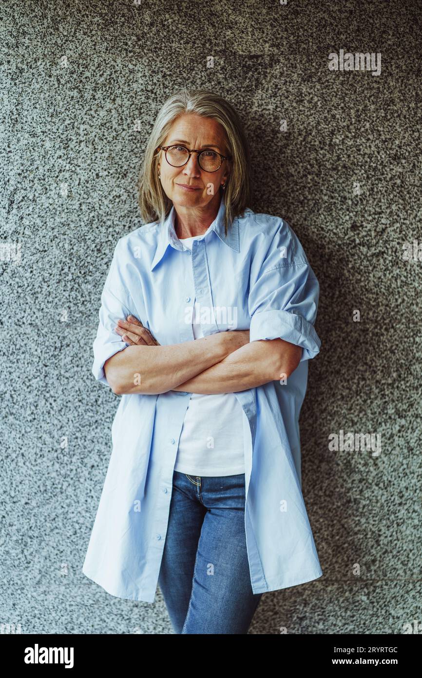 Reife Dame mit grauem Haar, die in der Nähe der Wand steht. Das selbstbewusste Verhalten der Dame spiegelt ihre Weisheit und Erfahrung wider. Sie strahlt Luft aus Stockfoto