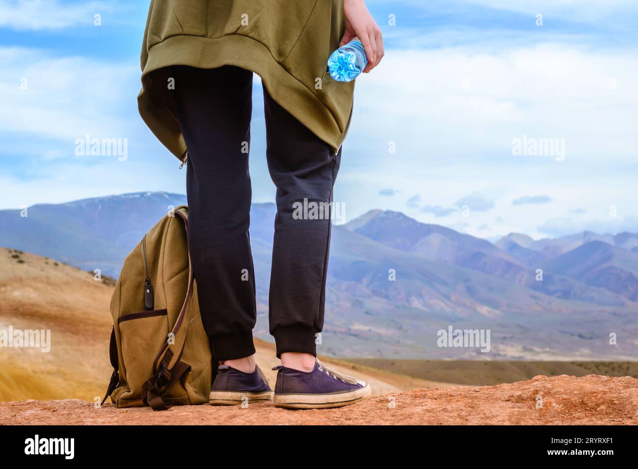 Mädchen-Tourist steht auf einem Hügel mit einer Flasche Trinkwasser in der Hand. Beine des Reisenden und Nahaufnahme des Rucksacks Stockfoto