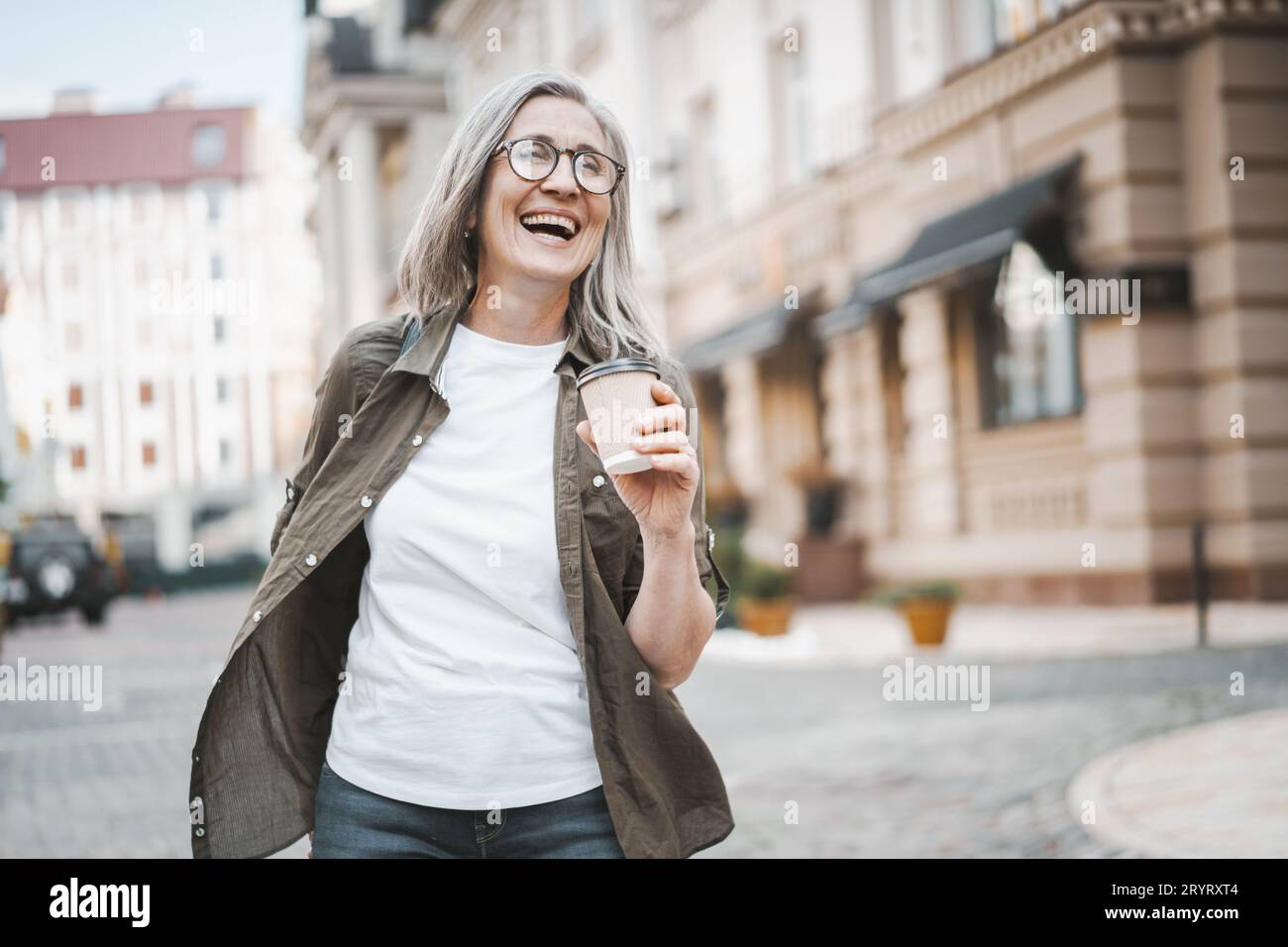 Das Konzept des glücklichen Alters durch das Bild einer silberhaarigen reiferen Seniorin, die in der Stadt spaziert und eine Tasse Kaffee genießt. Frau ist ca. Stockfoto